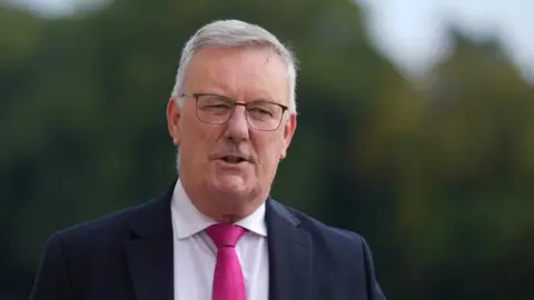PA Media A man with white hair and black rimmed, square glasses looks away from the camera as he talks. He is wearing a pink tie, white shirt and dark suit.