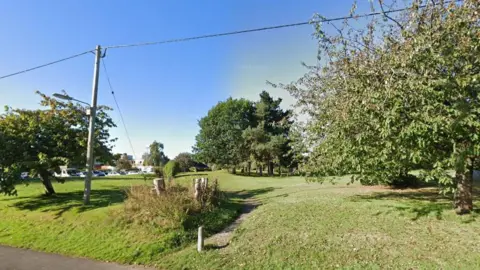 A screenshot from Google Street View of a park with green grass and trees, on a sunny day. The sky is blue.