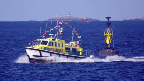 Tony Rive The Flying Christine is at sea and is dressed with a string of flags which extends from stern to bow. She has the words AMBULANCE along her port side and the reflective green and yellow ambulance style checker pattern. She has just passed a large marker buoy and in the distance there are islands, one of which has a beacon on it and another has a blue and white striped lighthouse on it.
