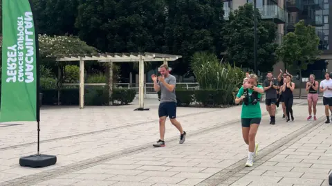 A group of runners with Daisy is approaching a finish area along a wide paved walkway in a residential complex near the water. Green vertical flags on either side display the text “Macmillan Cancer Support.” Spectators stand nearby clapping, including a child holding green and white pom-poms. Modern apartment buildings with glass balconies and landscaped greenery line the background.