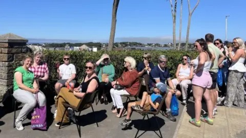 A queue of people sat outside a park with palm trees in the background.