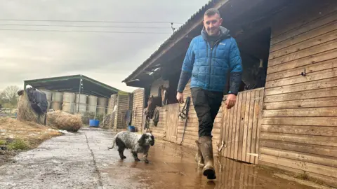 Farmer Andrew Jennings wears a blue jacket, black trousers and wellington boots as he walks through a farmyard next to stables, accompanied by a small black and white dog.