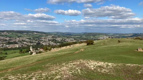 A sloping limestone hill in the Cotswolds, perched overlooking a valley with between 100 and 200 houses and a church. There are other hills surrounding the valley. It is mostly sunny day with some clouds in the sky.