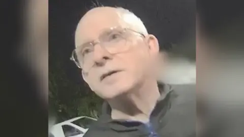 Sussex Police A man wearing a black quarter zip and glasses. He is standing in the dark.