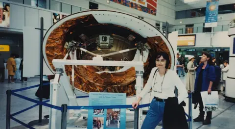 Michelle Dujeri Michelle Dujeriti is 30 years old at a white summit, blue jeans with glasses hanging around her neck and a black jacket flowing on her arm. It stands in front of a round part of the spacecraft that is partially white but with colored chips of copper in parts. It is cordoned out by the blue tape railway. The scene appears to be in a museum, or may wander the airport and other people.