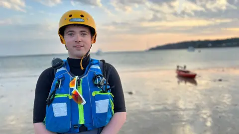Julian is wearing a yellow helmet and a blue life jacket stands on a beach with the ocean in the background. The sky is partly cloudy, and a small red boat is visible on the water near the shore