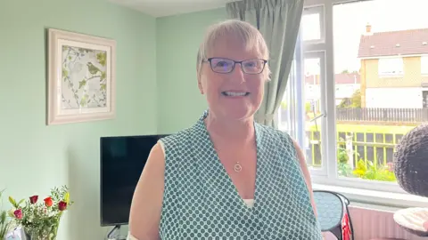 Woman standing in a living room with a window in the background as well as a TV, some flowers and a picture of a bird on the wall. She is smiling into the camera.