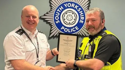 A tall bald smiling man in a white shirt hands a framed certificate to a police officer with grey hair and a beard, wearing a neon vest. They are both smiling at the camera.