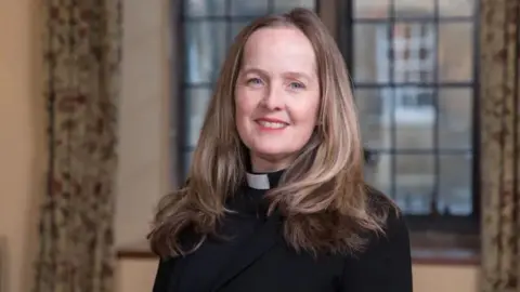 Jennifer Adams-Massmann is standing in front of a window in a room. She has long, wavy fair hair and is wearing a black robe and clerical collar