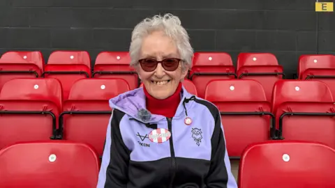 Amber Gash/BBC A woman sitting in one of the red seats at a football stadium. She has short grey hair and is smiling at the camera. She is wearing a black and purple zip-up hoodie with an Imps logo on it and two red and white pin badges. Underneath, she is wearing a bright red roll-neck jumper. She is also wearing sunglasses.