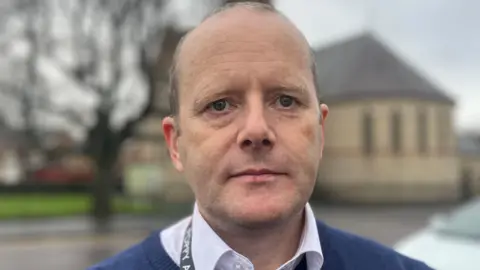 A man looking at the camera, he has light hair.