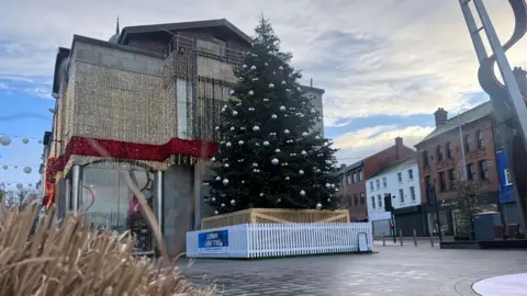 A large, real tree sits in Lisburn's Market Square. There are a number of silver baubles having from the branches, and the bottom of the tree is covered by both a pale, wooden box and a white picket fence. There are no people in the square, and it is a nice day.