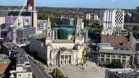Image shows an ariel shot of Leeds Civic Hall with Millenium Square. 
Behind is a Leeds University glass building with a sky line scene.
To the left of the shot is the Leeds General Infirmary.