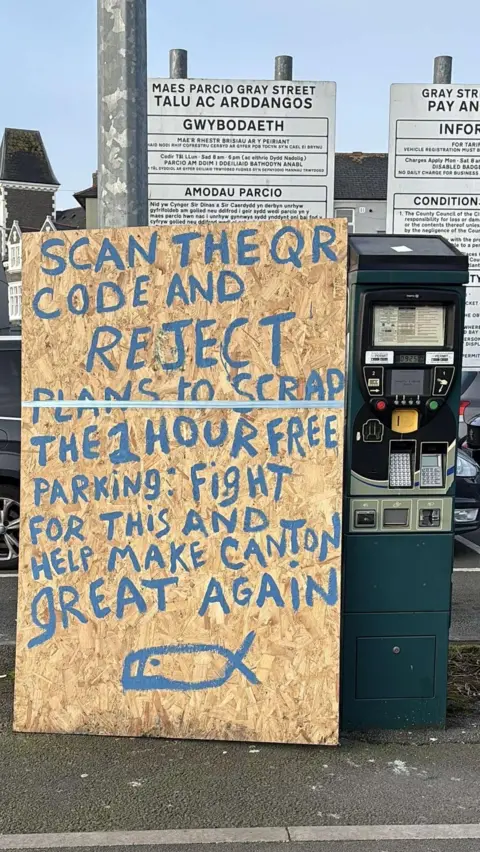 A large sign propped up against a parking meter in a Canton car park that reads "scan the QR code and reject plans to scrap the 1 hour free parking: fight for this and help make Canton great again'. 