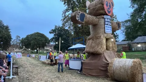 Giant straw bear with comical bear face in the foreground and people in hi viz jackets congregate around a wooden cubicle. There is a string of lights in the background.