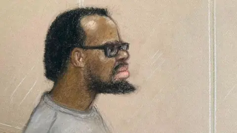Court sketch of Simon Levy, wearing a long sleeved grey top and thick rimmed glasses. He has short dark hair which appears to be receding, and a beard.
