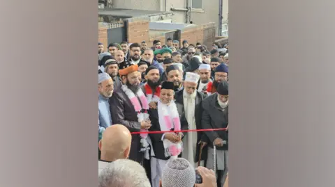 Tabassam Zabier A crowd of men smiling, waiting to cut a red ribbon 