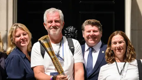 Two women and a man stand next to Mike McCarthy. He has short grey hair and a beard and is wearing a white T-shirt. He is holding a large gold baton. They are standing in front of the black door of 10 Downing Street.