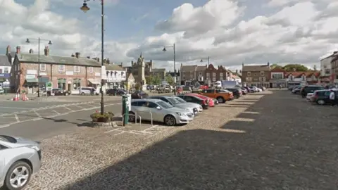 An Google streetview image of Thirsk Market Place. Two rows of cars are parked on a cobbled surface surrounded bu buildings.