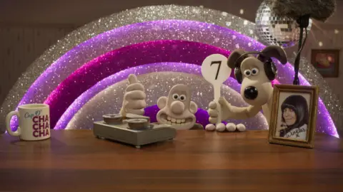 Aardman/BBC Wallace and Gromit still from guest appearance during Strictly's semi-final show. Sparkly silver and purple rainbow backdrop, disco ball on the ceiling in the top right of the picture. Wallace is peering over the top of a desk with a thumbs up. Gromit is wearing headphones, and in one hand holds a mic and in the other, a score paddle with a "7" on it. On the desk, is a framed Claudia Winkleman autographed photo, a tape player and a mug that reads "cup o' cha cha cha".