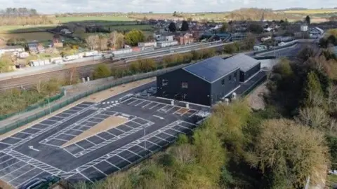 The building resembles two large black sheds, one much bigger than the other one. On the left of the photograph, which is taken from the air, a new carpark has been marked out. The building is surrounded by trees and the village can be seen on in the distance.