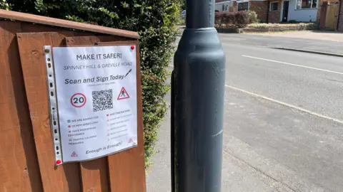Image of a fence near a roadside with part of a street light in the foreground. There is a hedge above the fence, on the fence is an A4 sign in a plastic wallet it is a petition to call for drivers to drive through the area more safely.
