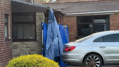 A blue tarpaulin and a blue frame covers an entrance to a home, with a silver car parked close to it