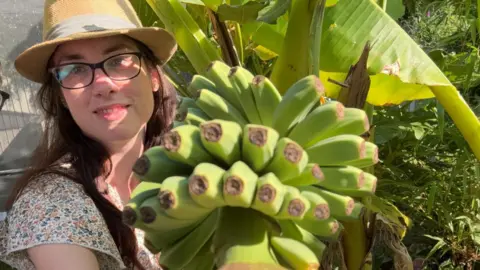 Supplied A woman with glasses and a hat posing next to a bunch of bananas