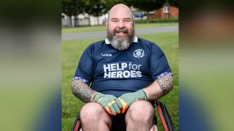 Dave Anderson sat in a wheelchair. He is smiling at the camera. Dave is wearing a blue Help for Heroes T-shirt and has tattoos on both arms.