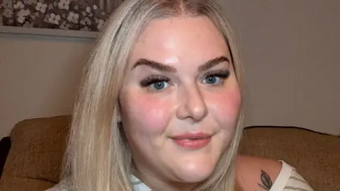 A woman with blonde hair smiles with closed lips as she looks at the camera. She is sitting on a beige sofa in a room and has a striped green and white top slipping off her shoulders