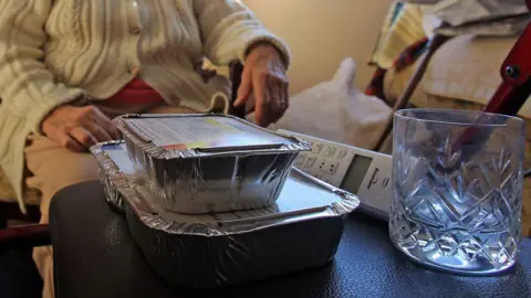 Two foil food containers stacked, and empty glass and a landline wireless phone sit on a black leather-like surface. Behind it is an elderly person wearing a beige cable knit cardigan with buttons. Face is not visible and clothing does not make it possible to distinguish if they are male or female.