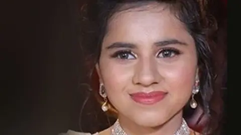 A close up photo of Harshita Brella wearing a white dress, a diamond looking necklace and gold earrings and red lipstick. She is smiling without teeth