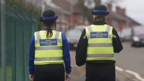 Two people in high-visibility vests with the words Police Community Support Officer on the back are walking away from the camera, on a residential street.