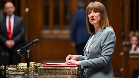A woman wearing a green/grey jacket stands in the House of Commons and looks to her left as she gives a speech. Her hands are resting on a red folder. Blurred figures can be seen watching in the background.