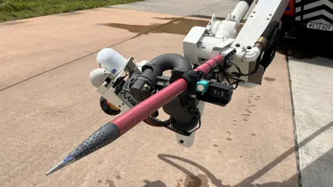 A large, metal spike is attached to the front of a fire engine. The tip is punctured with holes - for spraying water or foam. Its shaft is red - and the tip is grey and silver. Several wires are connected to it.