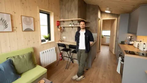 A man - John Littlejohn - standing indoors, in a small modular home. There are various chairs, tables, a kitchen counter and shelves. The majority are wood panelled.
