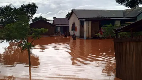 An image of a town in Madagascar flooded with dark muddy water. The buildings are clearly severely flooded. There are people wading through the water. 
