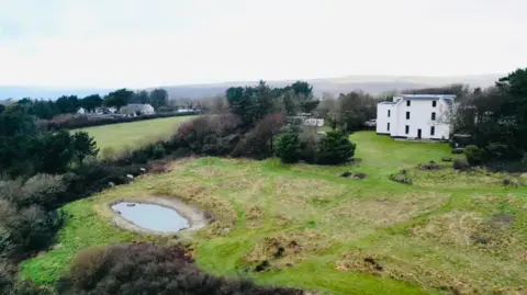 A drone shot shows the large white £1.1m house at Berry Park in the background with an expanse of land in front and a large pond in the foreground, surrounded by trees and grass. To the left in the distance can be seen about five other properties in a cluster. 