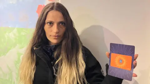 An image of a woman wearing a black coat, standing in front of a white wall, while holding an Aldi voucher. She is looking straight into the camera.