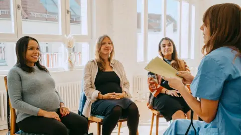 Getty Images A small prenatal information session where three pregnant women sit in a bright room while a healthcare professional leads a discussion. The atmosphere feels calm and supportive, with everyone focused and engaged.