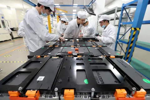 AFP via Getty Images A Chinese factory with batteries open on a work table, workers in white lab coats and wearing blue masks are gathered round the batteries. The frames of the building are painted yellow and blue, with yellow and black warning tape on them. 