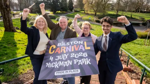 Two women and two men smile and raise their arms while they stand in a park holding a large purple sign which says "Bilston Carnival, 4 July 2026, Hickman Park".