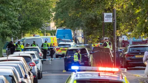 An area of a street with cars parked all along the left side is cordoned off with police tape. At least eight uniformed officers all stand around the cordon while a number of police cars and vans are in the background. Flashing police lights are in the foreground.