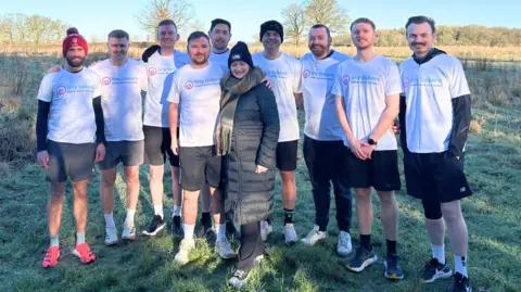 Steve has his arm around Ruth's shoulder. Steve and the team wear black shorts and white tops. Ruth wears a black puffer jacket with a hat and scarf.