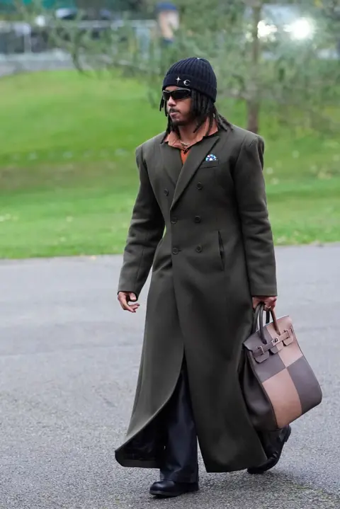 Getty Images Picture of Jules Kounde in a long green jacket, holding a handbag and wearing a beanie. There is grass and concrete behind him.