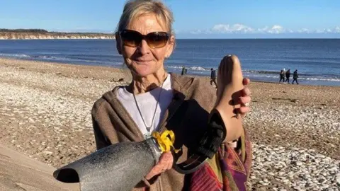 Brenda holds the prosthetic up - she is on the beach smiling. The sun is out and she is wearing sun glasses.