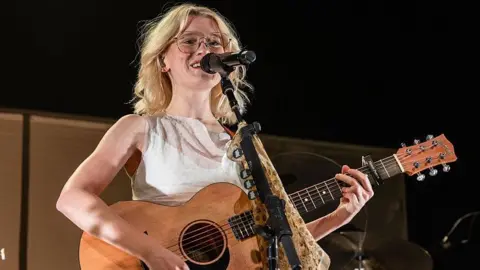Getty Images Maisie Peters plays a guitar as she performs on stage with her band in the background. The singers has short blonde hair cut at shoulder length, with large glasses and wears a white linen top. 