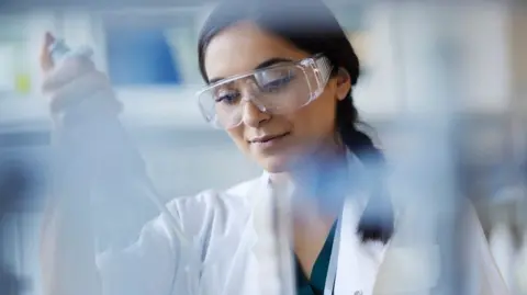 A young scientist in safety goggles and a white lab coat works in a bright laboratory. She concentrates on using a pipette to transfer liquid between clear containers, surrounded by softly blurred glassware and equipment, creating a focused, professional atmosphere.