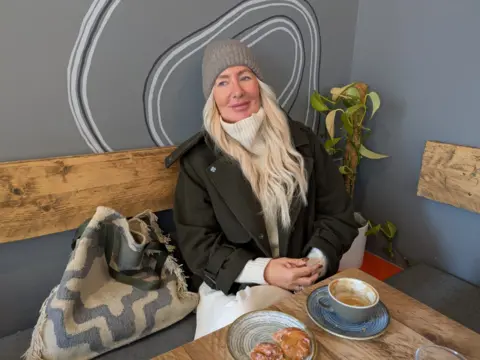 Karen Pearson se sienta frente a una masa y café en una cafetería independiente, con un sombrero de lanuda y una chaqueta