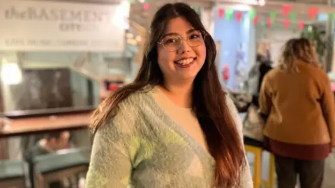 BBC/Seb Cheer A woman with long brown hair and wearing glasses smiles at the camera. There is pink and green bunting behind her.